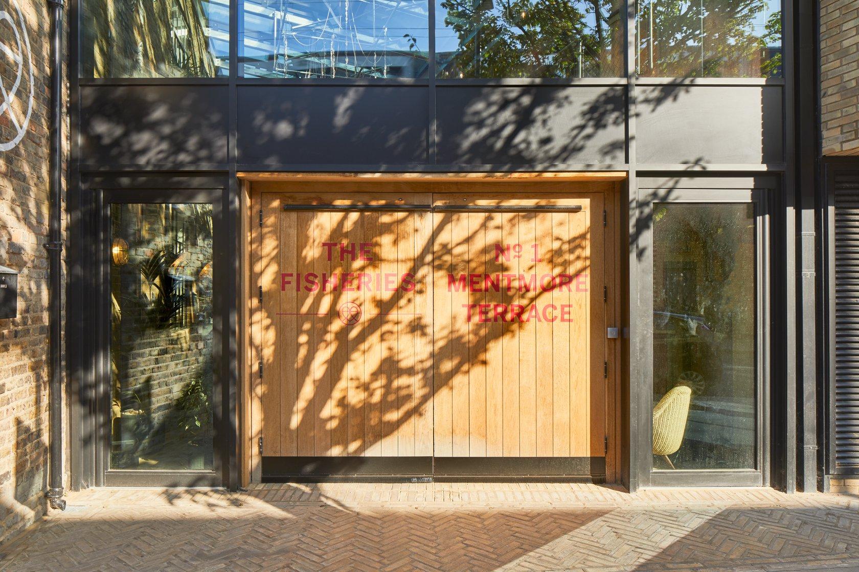 A coworking space with a wooden entrance door, large glass windows, and brick facade at The Fisheries.