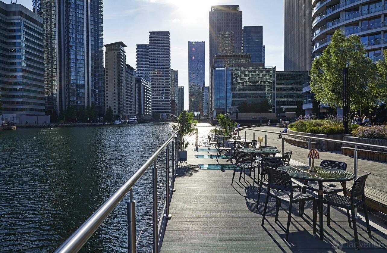 A waterside bar on a boat with metal tables and chairs along the deck overlooking Canary Wharf at The Boathouse London.