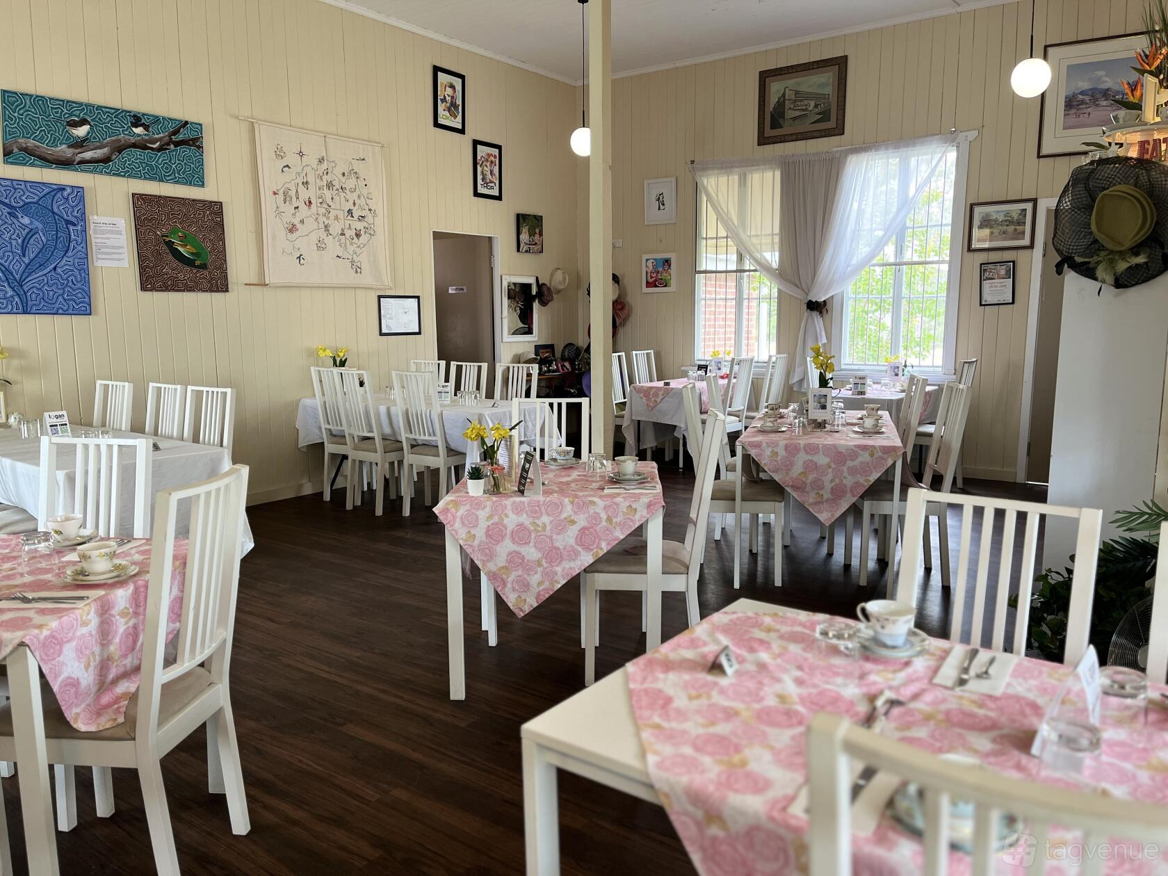 A cafe with pink floral tablecloths, white wooden chairs, and large bright windows at Devon Pixies.