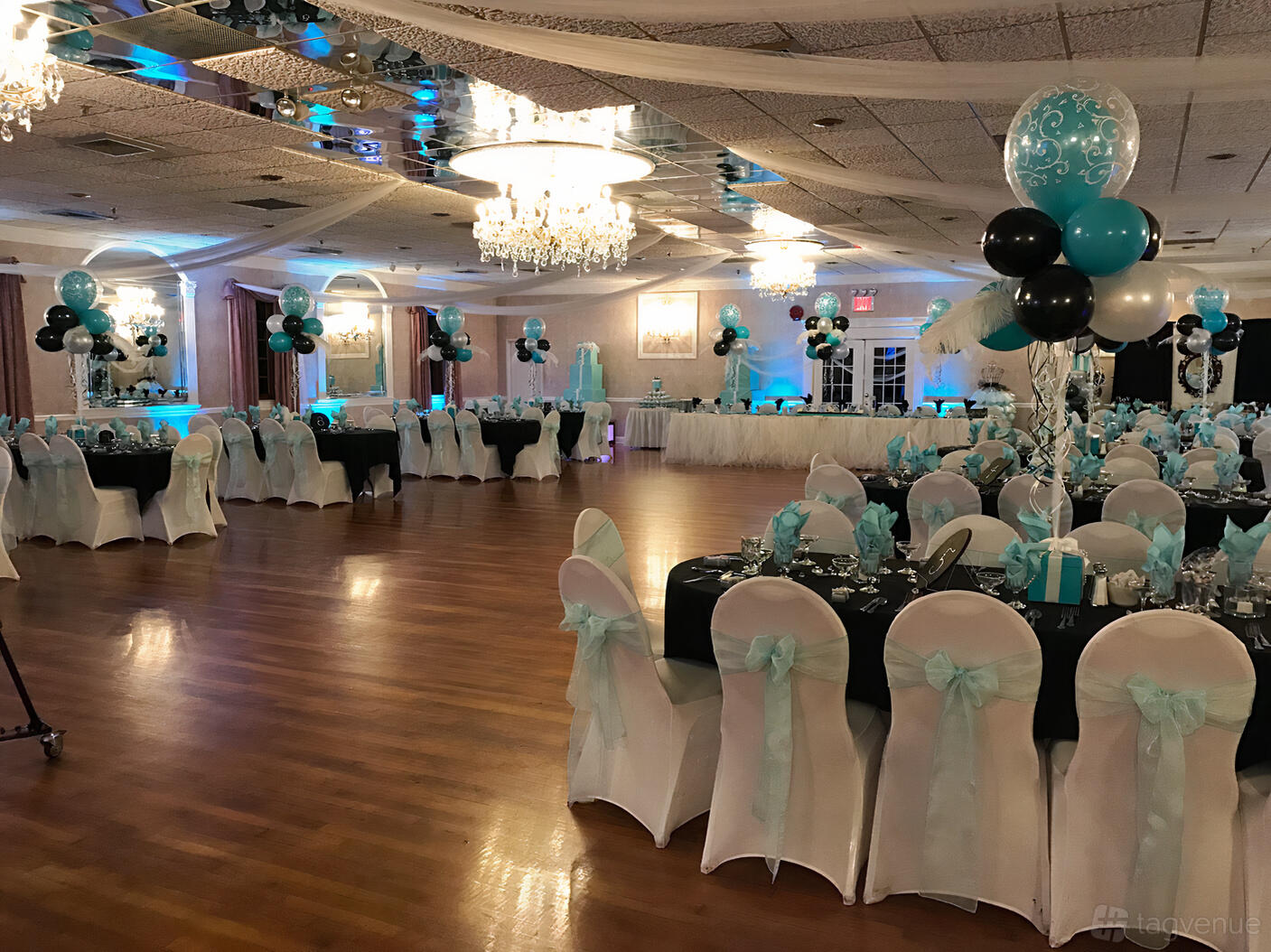 An event venue with chandelier lighting, hardwood floors, and round tables decorated with blue and white balloons at The Crystal Room.