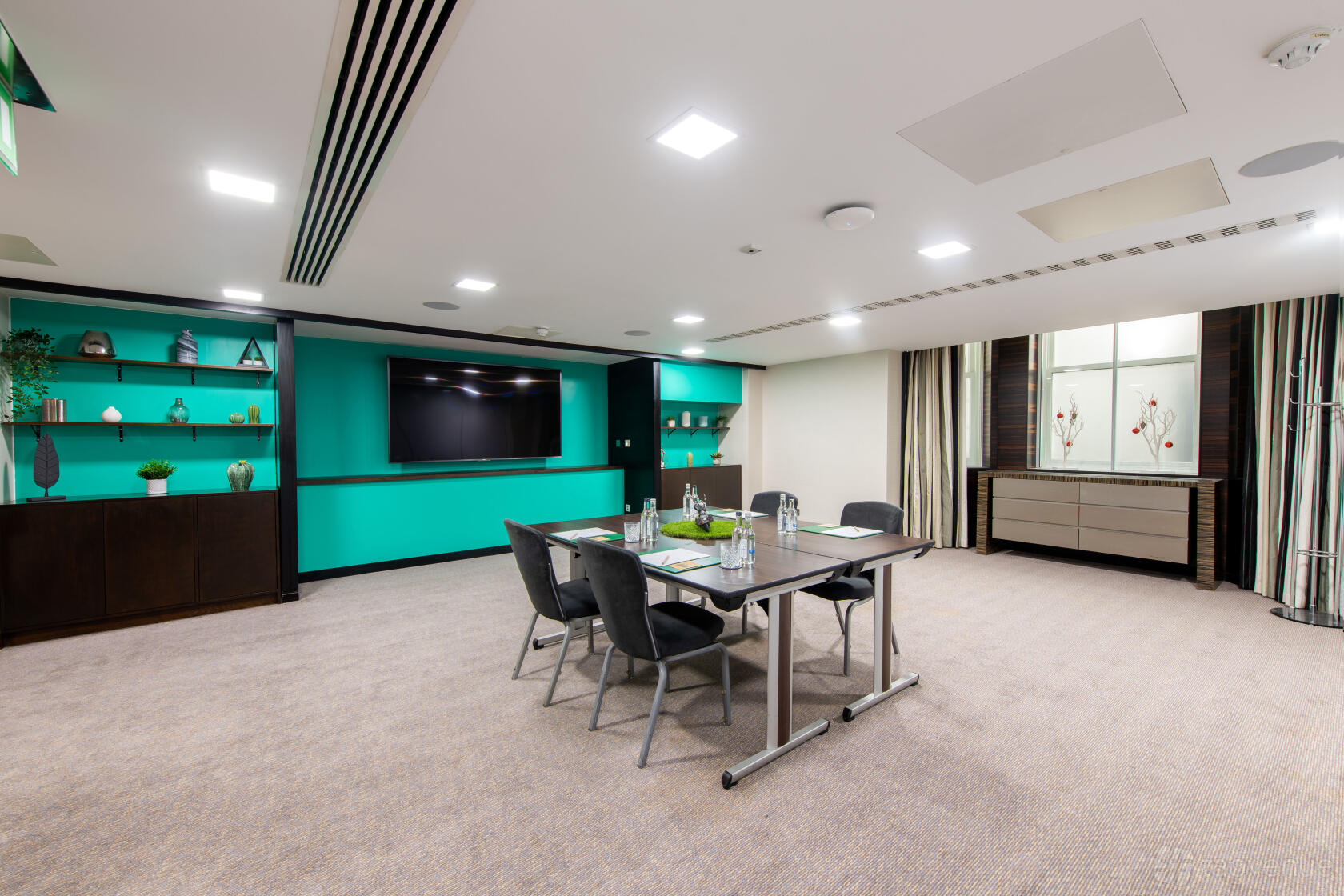 A hotel meeting room with teal accent shelves, a wall-mounted TV, and a table set for four at DoubleTree by Hilton London Kensington.
