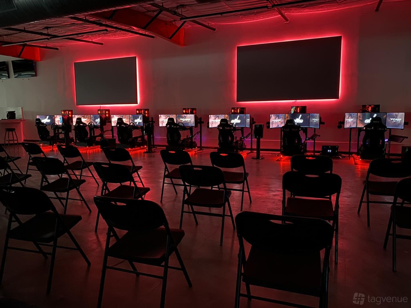 A warehouse space with rows of gaming stations, red LED lighting, and black folding chairs at The Asylum.