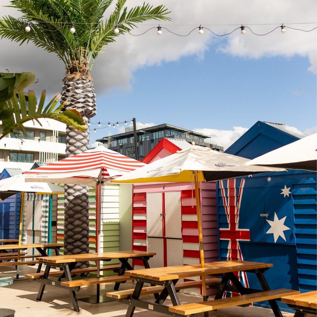 An outdoor bar space with colorful beach boxes, picnic tables, and umbrellas at The Bungalow Beer Garden.