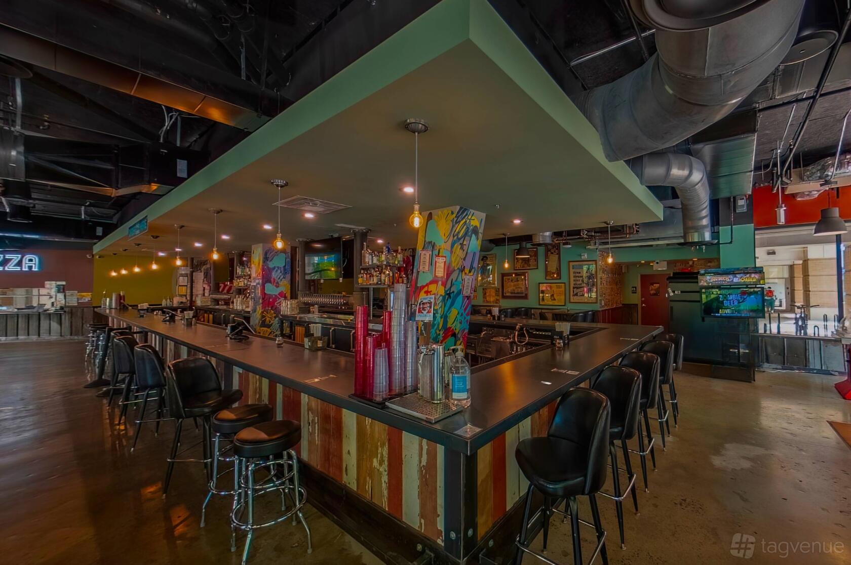 A restaurant dining room with a large central bar, colorful artwork, and industrial ceiling at Quincy Hall.