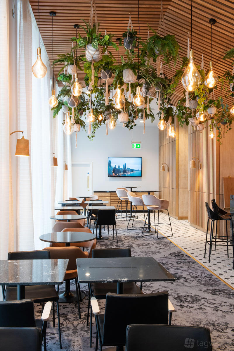 A hotel bar with hanging pendant lights, greenery, velvet bar stools, and floor-to-ceiling windows at voco Manchester.