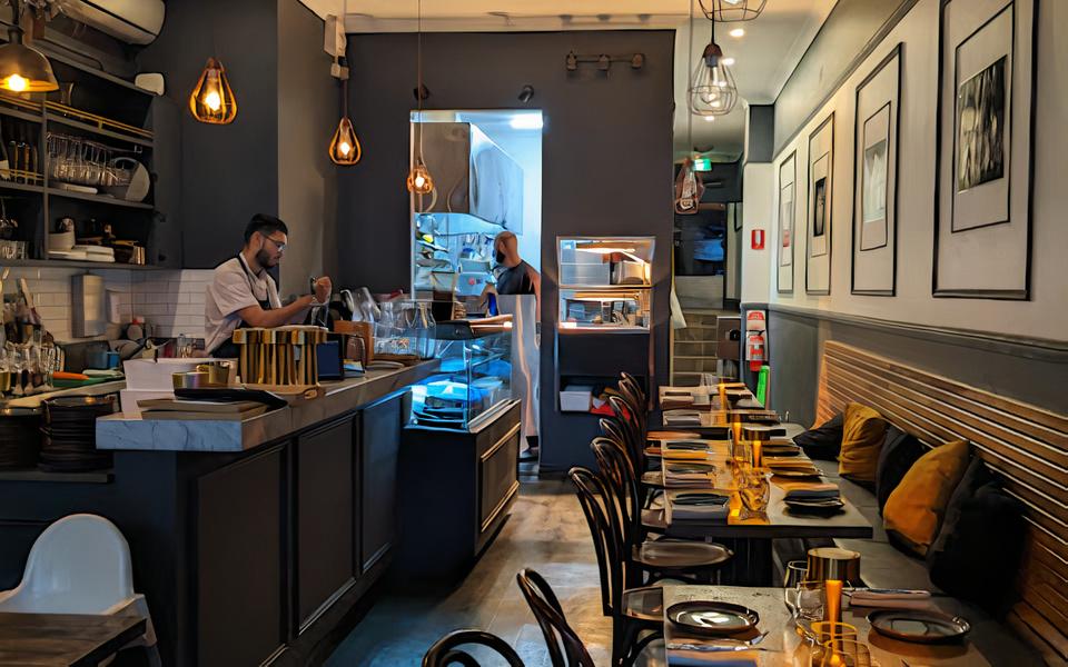 A restaurant with pendant lighting, dark wood tables set with gold-accented plates, and an open kitchen at Semola Sydney.