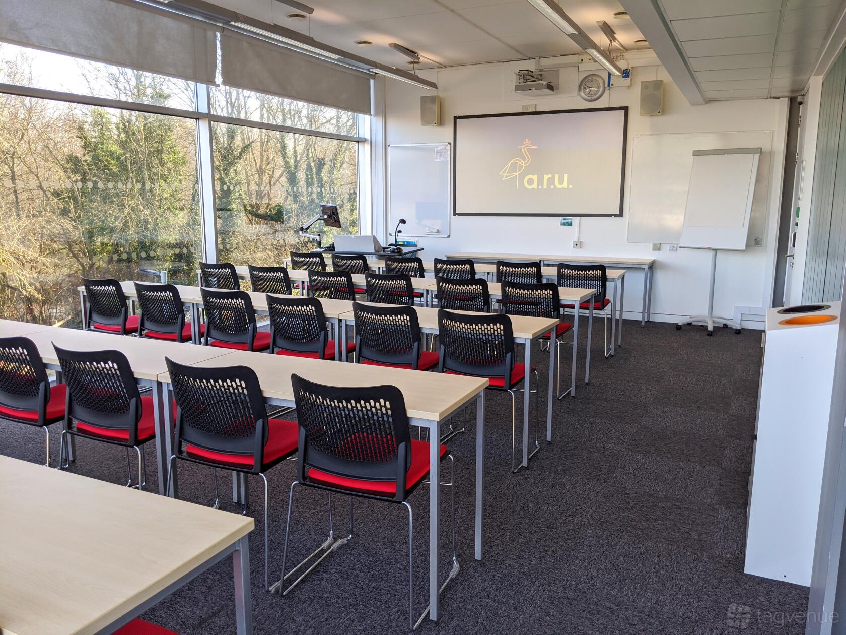 A classroom with rows of desks and black chairs facing a projector screen and large windows at ARU Venue Hire - Chelmsford.