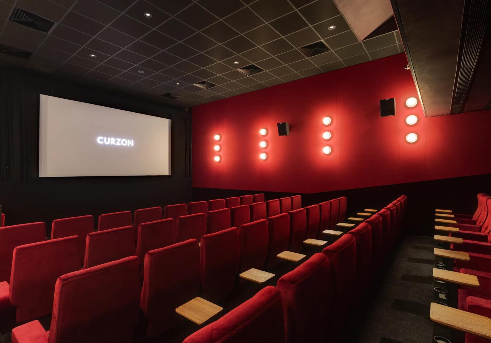 A cinema with red velvet seats, wall-mounted lights, and a large projection screen at Curzon Soho.