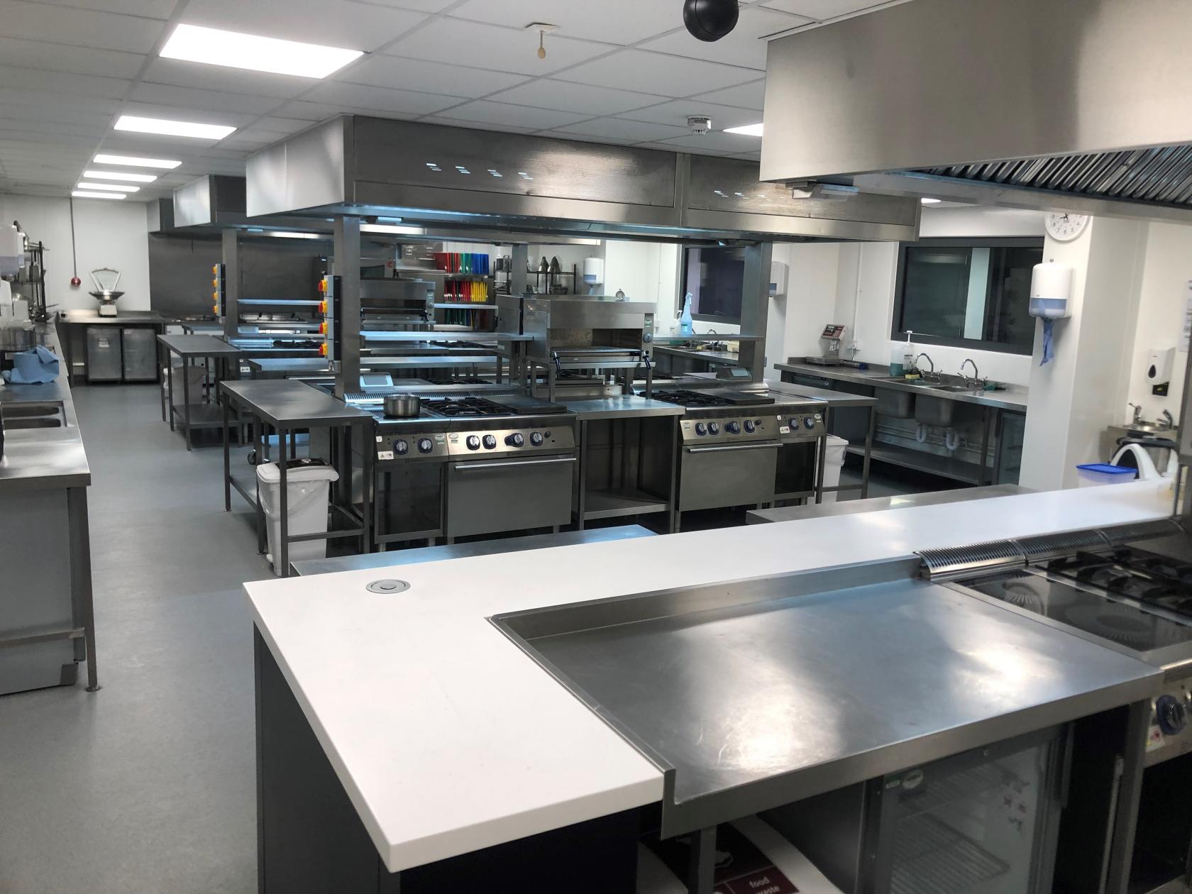A culinary classroom with stainless steel cooking stations, ovens, and open shelving at University of West London - Ealing Site.