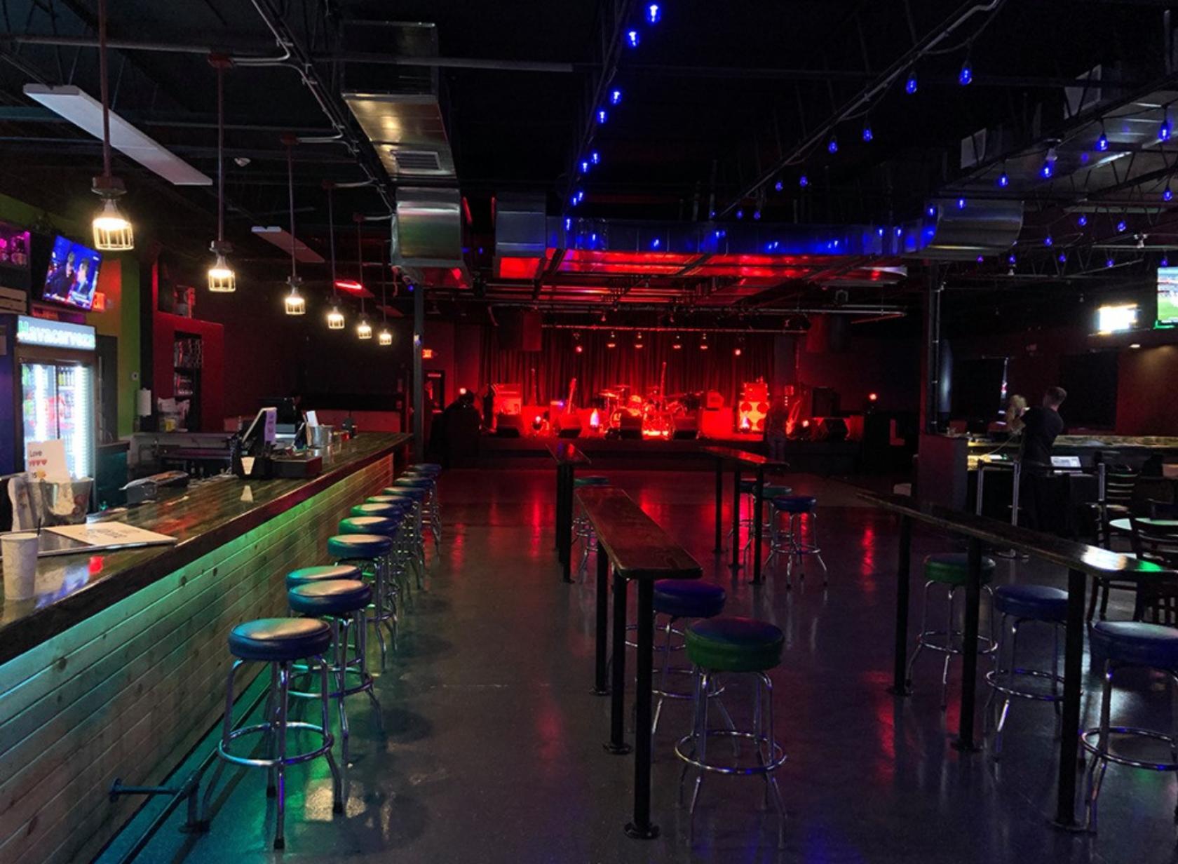 A nightclub with a long bar, high tables, barstools, and a stage lit with red and blue lights at The WC Social Club.