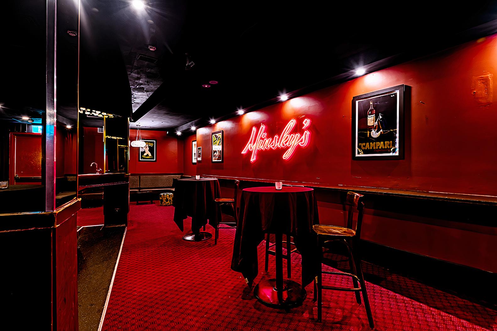 An event space in a pub with red walls, framed posters, cocktail tables, and neon signage at Minskys Hotel Piano Bar.