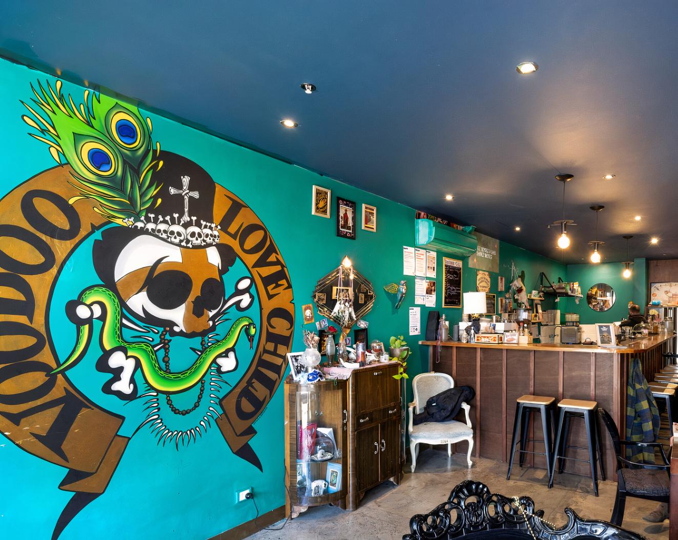 A bar with teal walls featuring bold mural art, a wooden counter with stools, and eclectic decor at Voodoo Love Child Speakeasy.