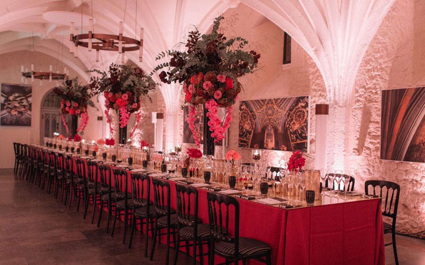 A dining room with vaulted stone ceilings, chandeliers, and long banquet tables with tall floral centerpieces at Westminster Abbey.