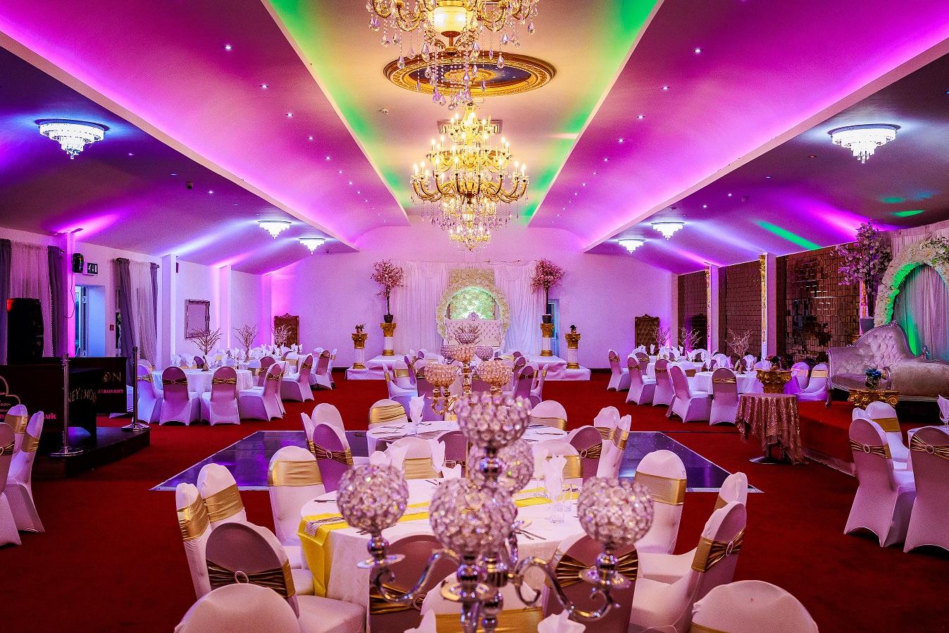 An event venue with crystal chandeliers, purple ceiling lights, and round tables with white and gold chairs at Honeymoon Banqueting.