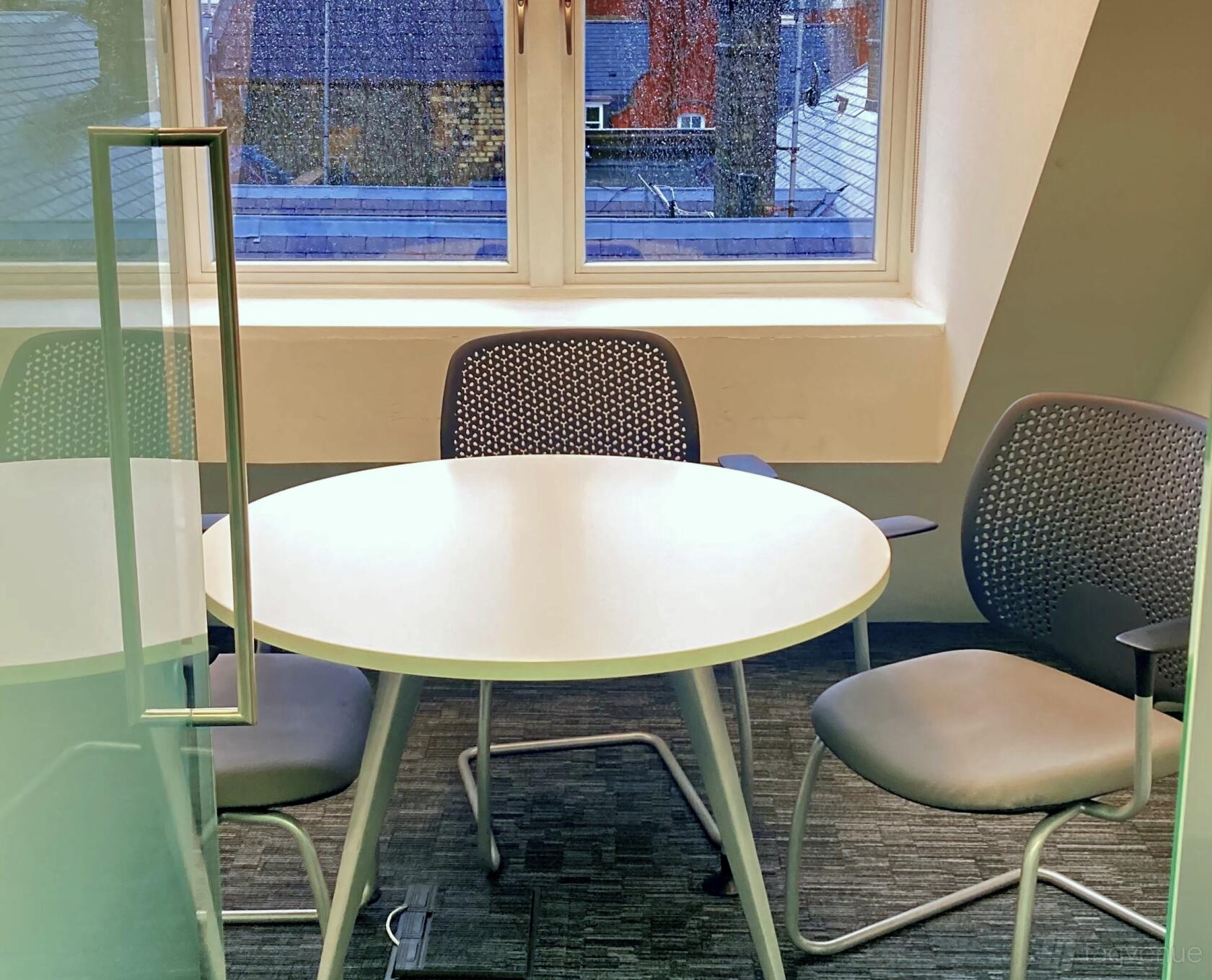 A meeting room with a round white table, three mesh chairs, and a window with rooftop views at Rivvia Pronto Meeting Rooms.