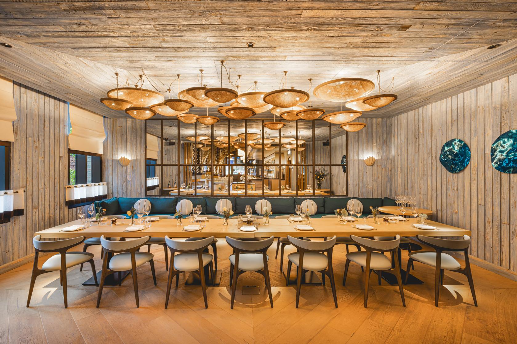 A dining room in a restaurant with a long table, modern chairs, wood paneling, and sculptural ceiling lights at Azzurra.