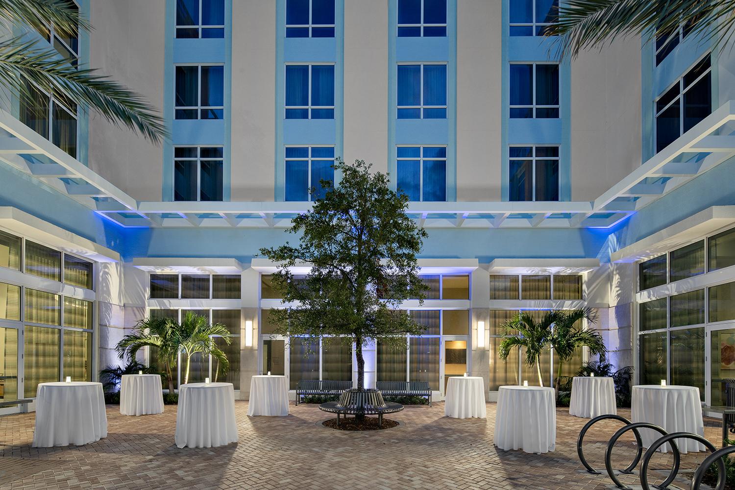 An outdoor courtyard with a central tree, palm plants, and cocktail tables with white linens at Courtyard Delray Beach.