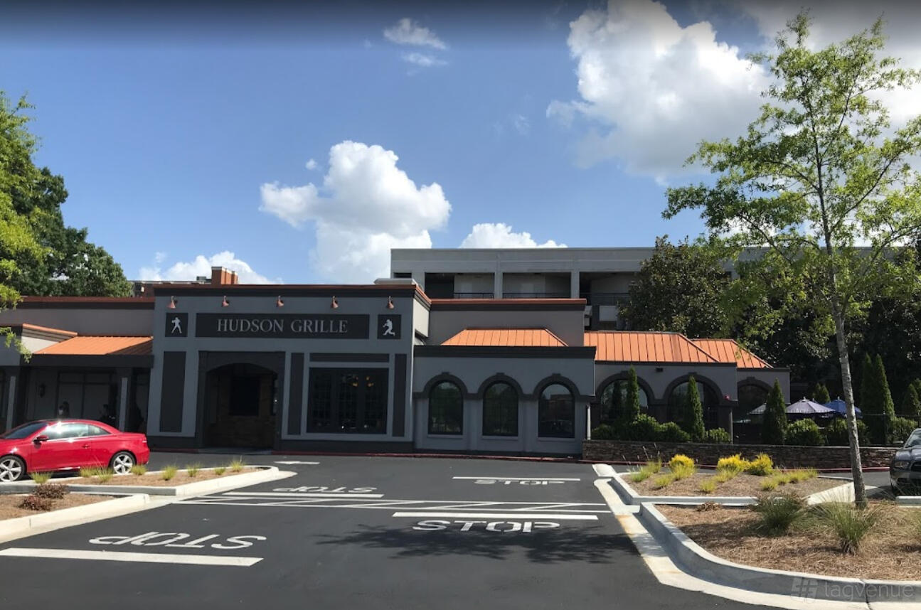 A pub with a gray facade, orange roof accents, arched windows, and front parking lot at Hudson Grille - Sandy Springs.