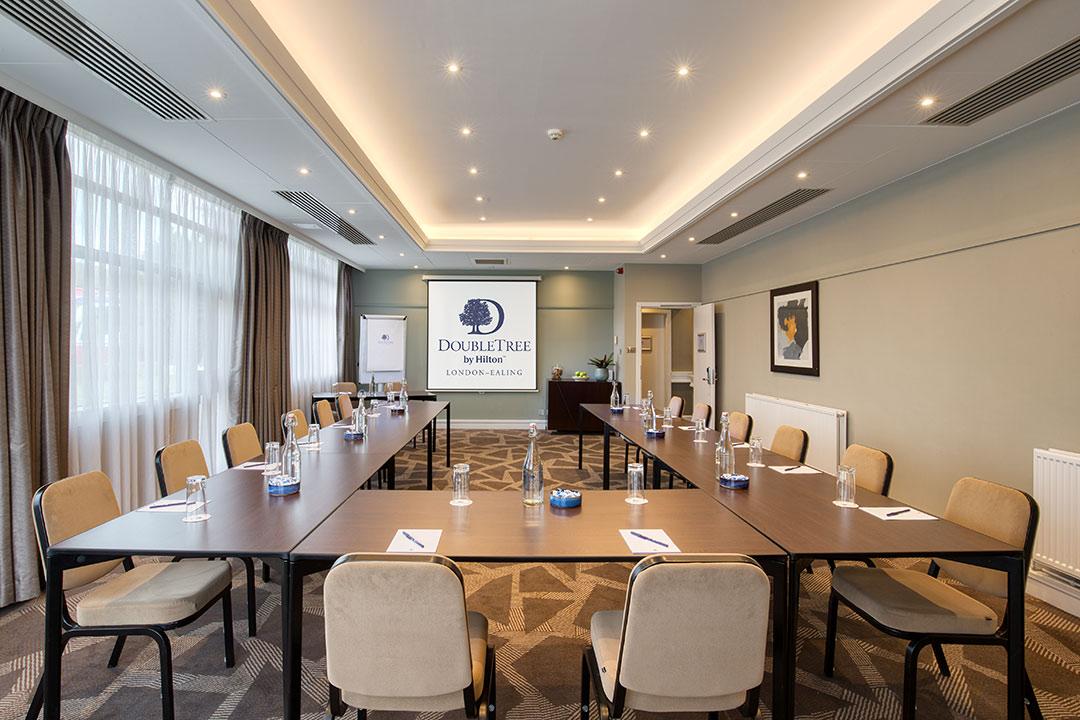 A hotel meeting room with U-shaped tables, beige chairs, carpeted floors, and ceiling spotlights at DoubleTree by Hilton London - Ealing.