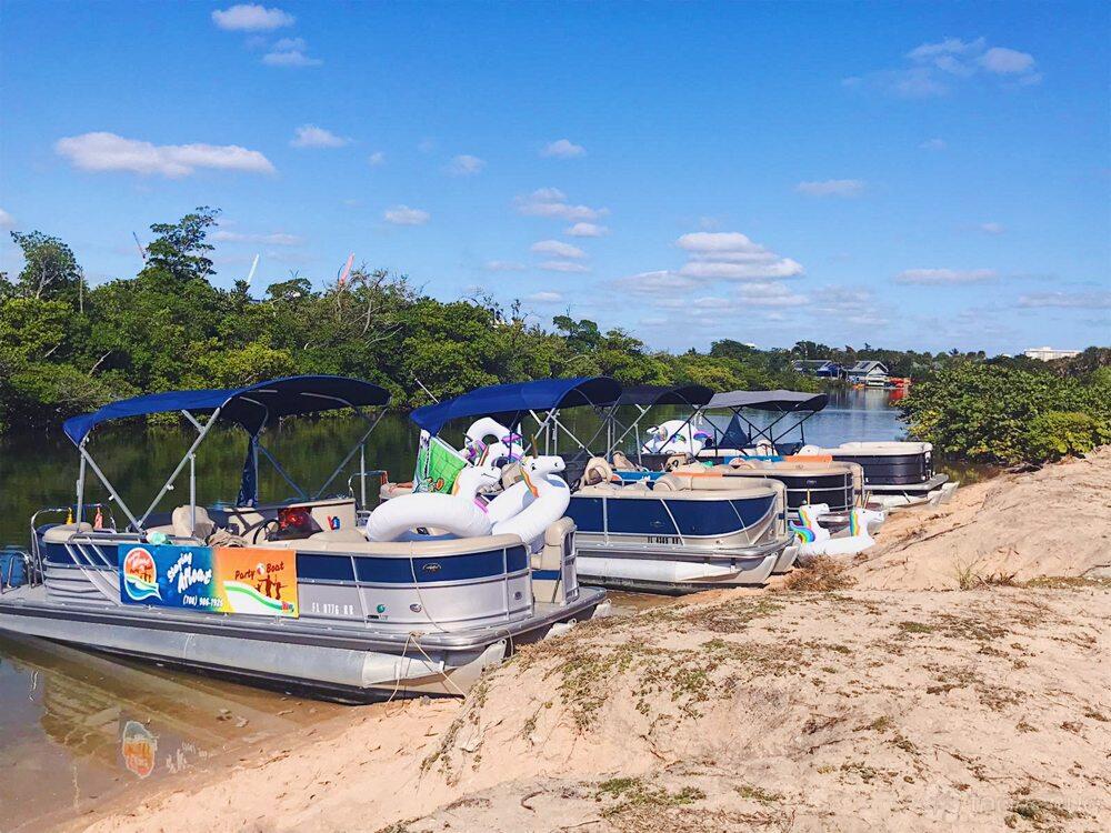 A fleet of pontoon boats with blue canopies and inflatable floaties docked along a sandy shore at Staying Afloat Boats.
