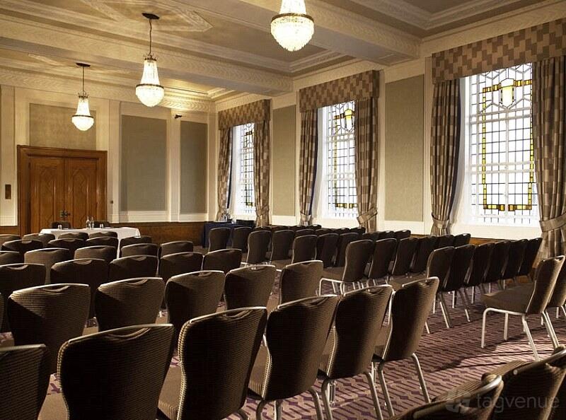 A conference centre with rows of black chairs, high ceilings, chandeliers, and tall windows at Grand Connaught Rooms.