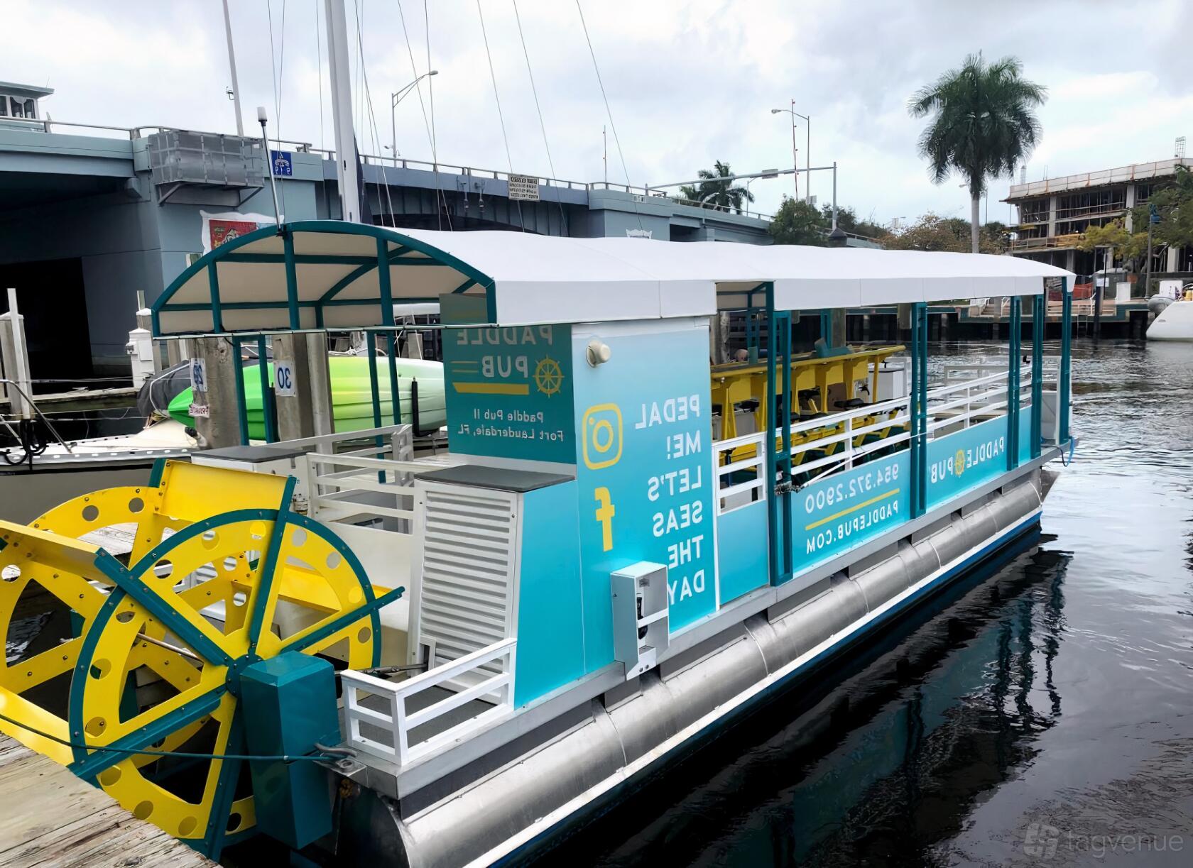 A party boat with covered seating, a rear yellow paddle wheel, and open sides at Houston Pedal Barge.