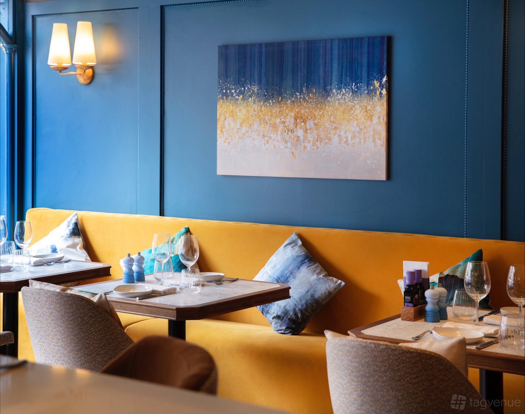 A restaurant with a yellow upholstered bench, blue accent wall, abstract art, and set tables at Bellazul Restaurant & Bar.
