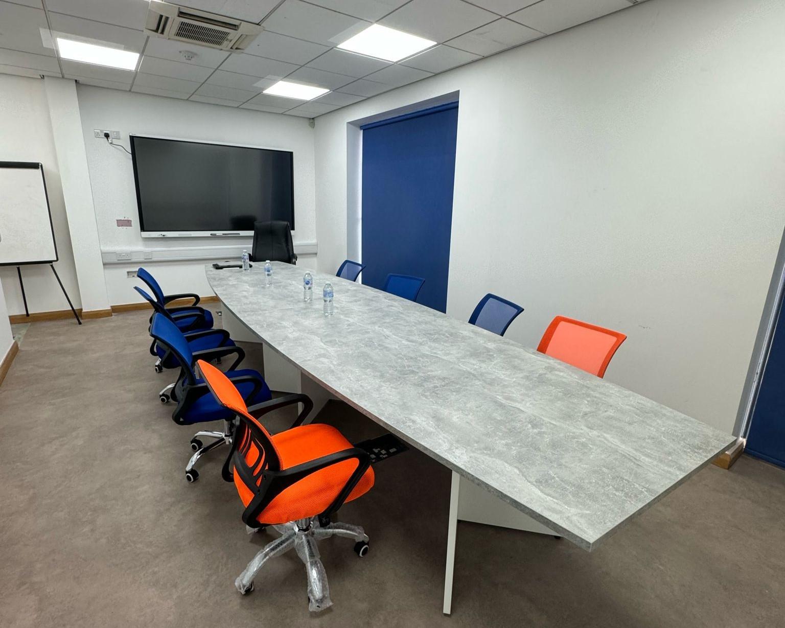 A conference room with a long table, colorful office chairs, a wall-mounted screen, and a whiteboard at MediCenter Bolton.