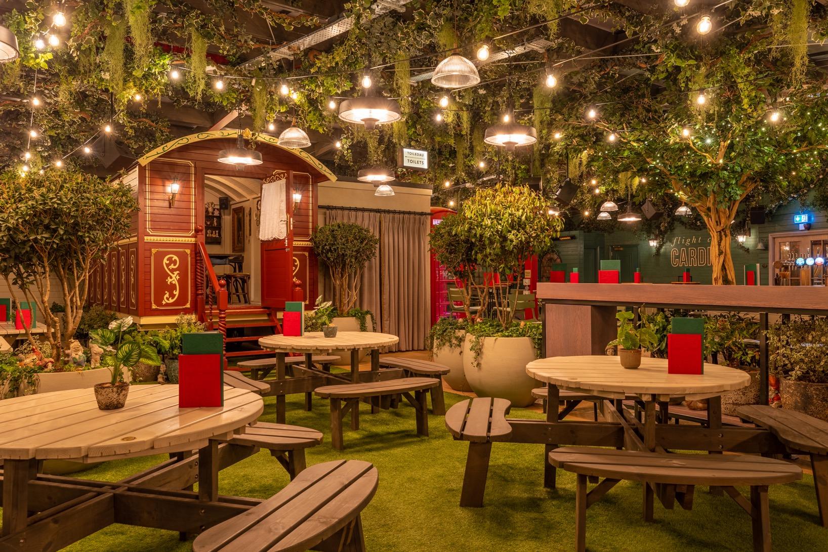 A bar with round wooden picnic tables, artificial grass, potted plants, and string lights at Flight Club Cardiff.