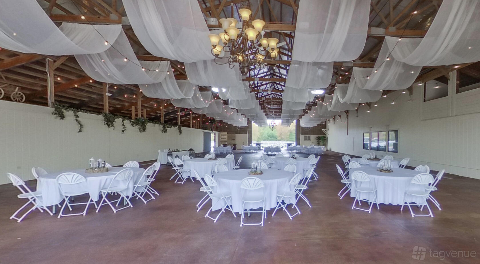 An event hall with round tables covered in white linens, draped fabric ceiling, and a central chandelier at BrighterDaze.