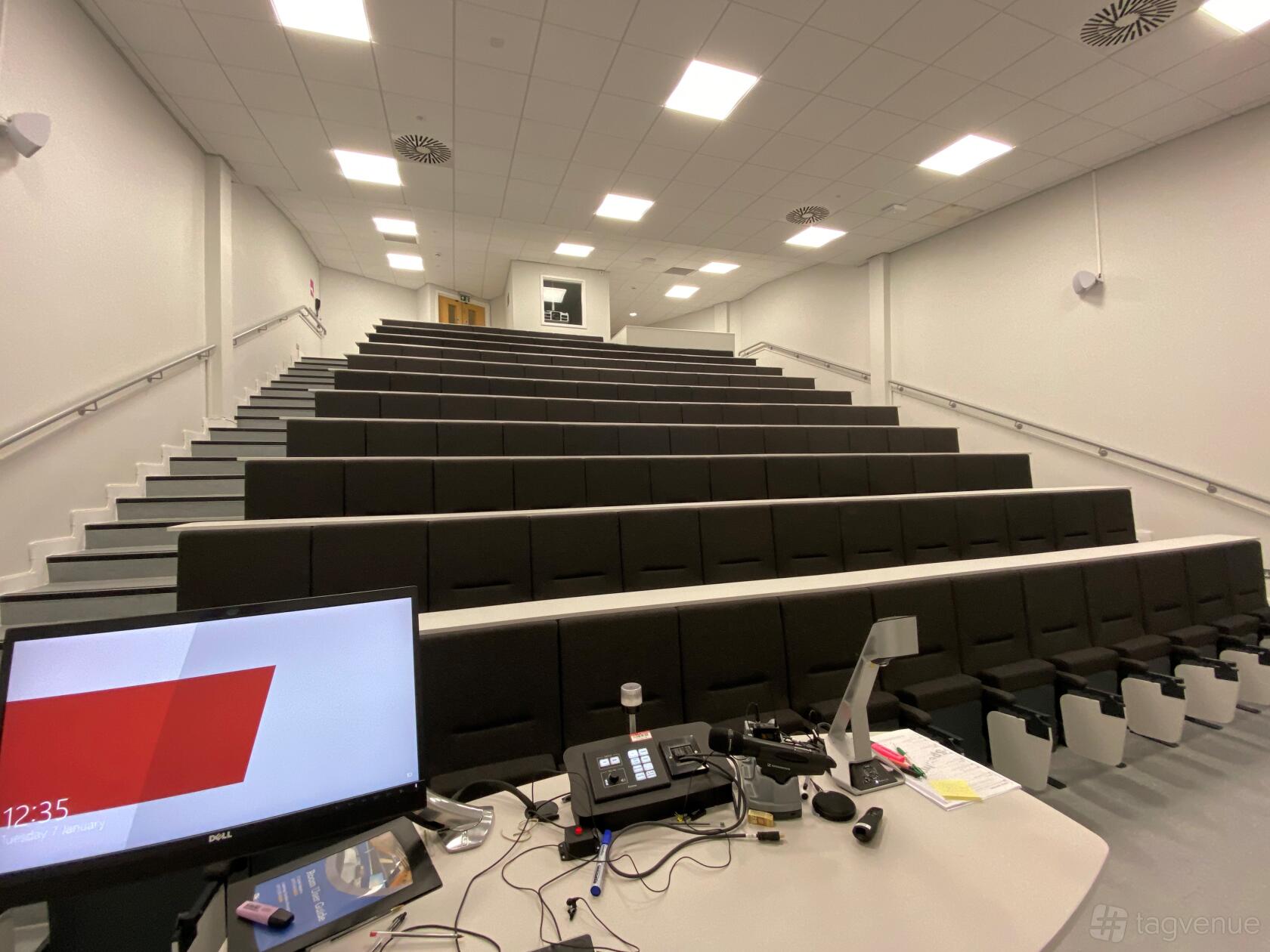 A university lecture theatre with tiered black seating, white walls, and a desk with AV equipment at Priory Road - Social Sciences Complex.