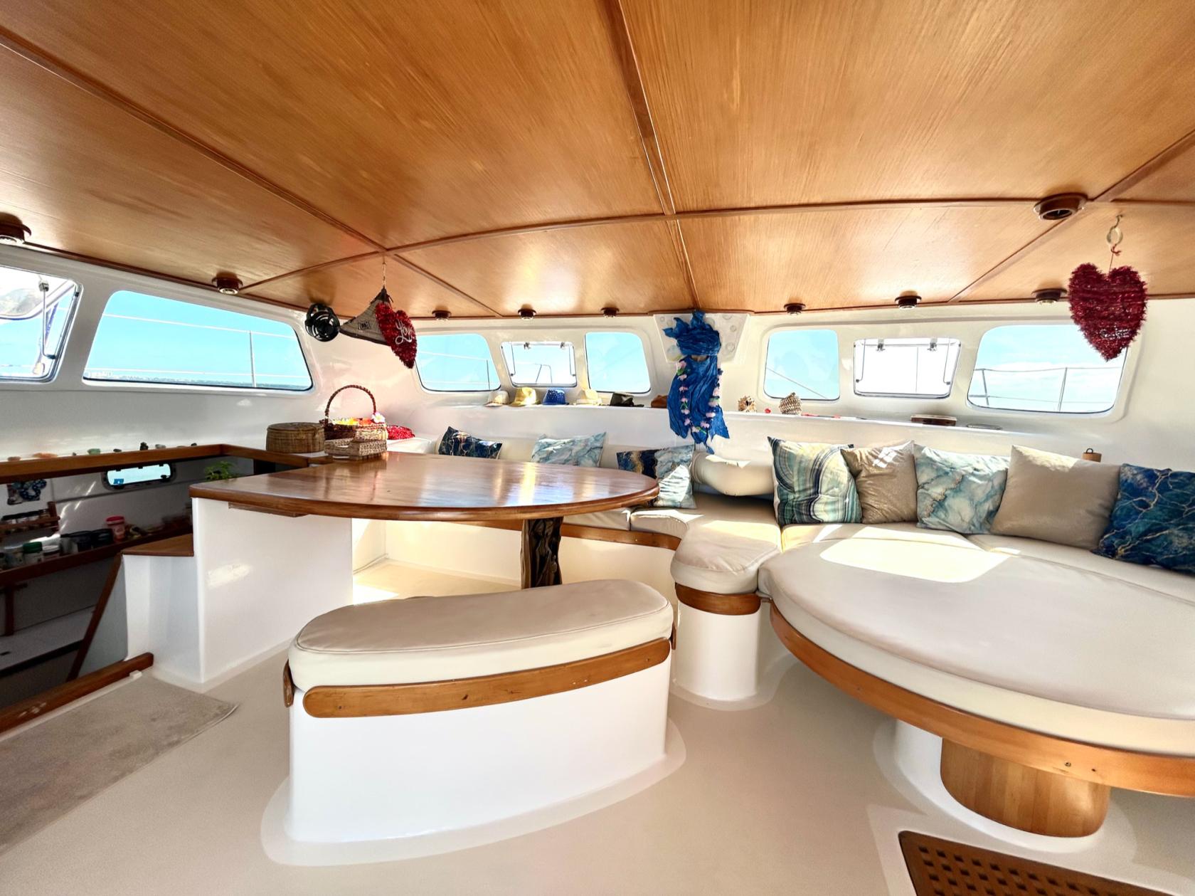 A boat interior with a round wooden table, curved cushioned seating, and large windows on Sailing Bubble.