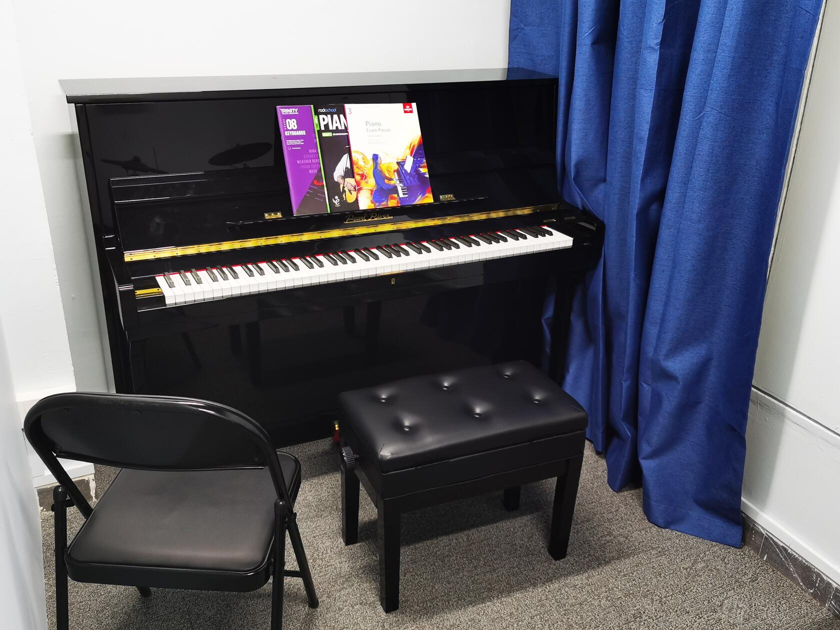 A piano room with an upright piano, padded bench, and folding chair beside blue curtains at Anvil Music Productions.