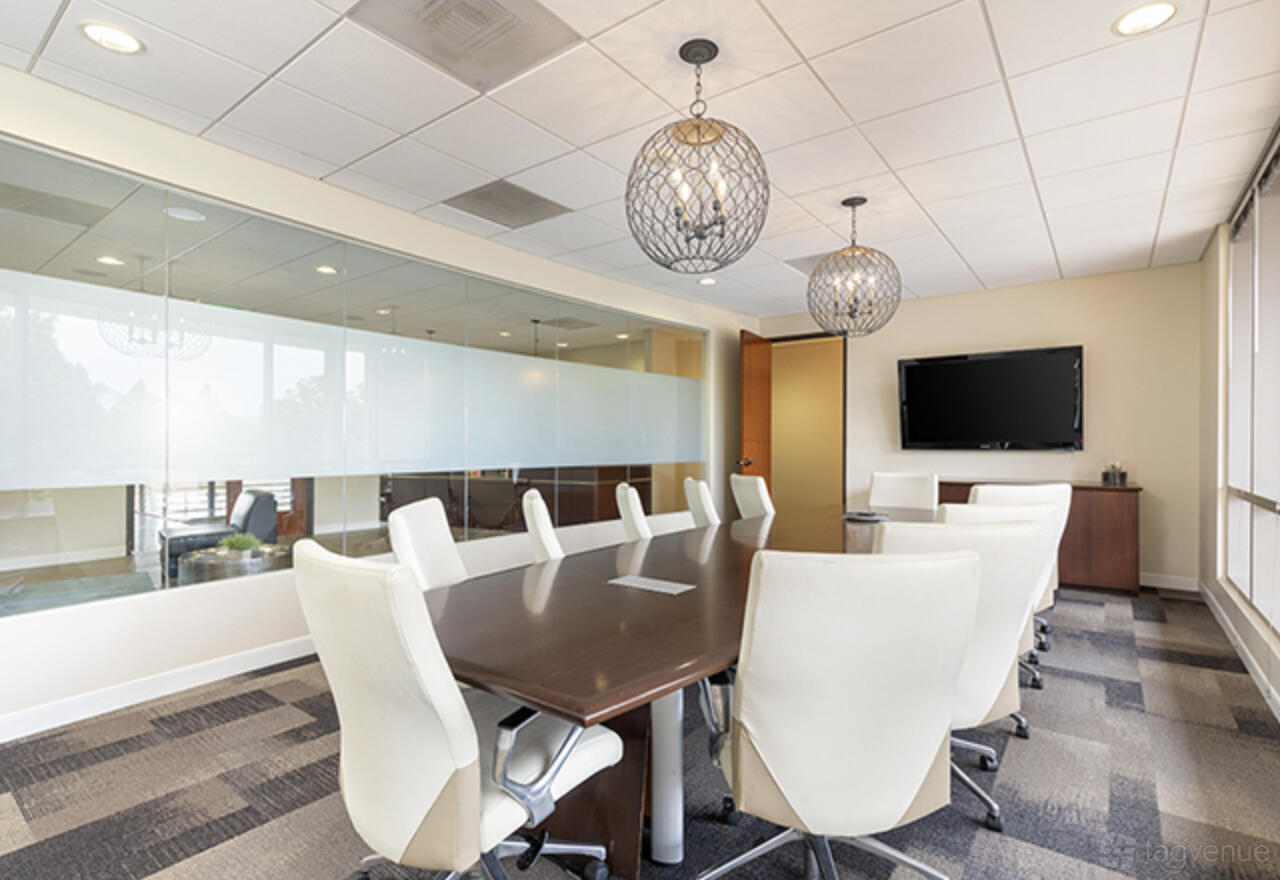 A coworking space with a long conference table, white chairs, pendant lights, and a wall-mounted TV at Premier Workspaces - Carlsbad.