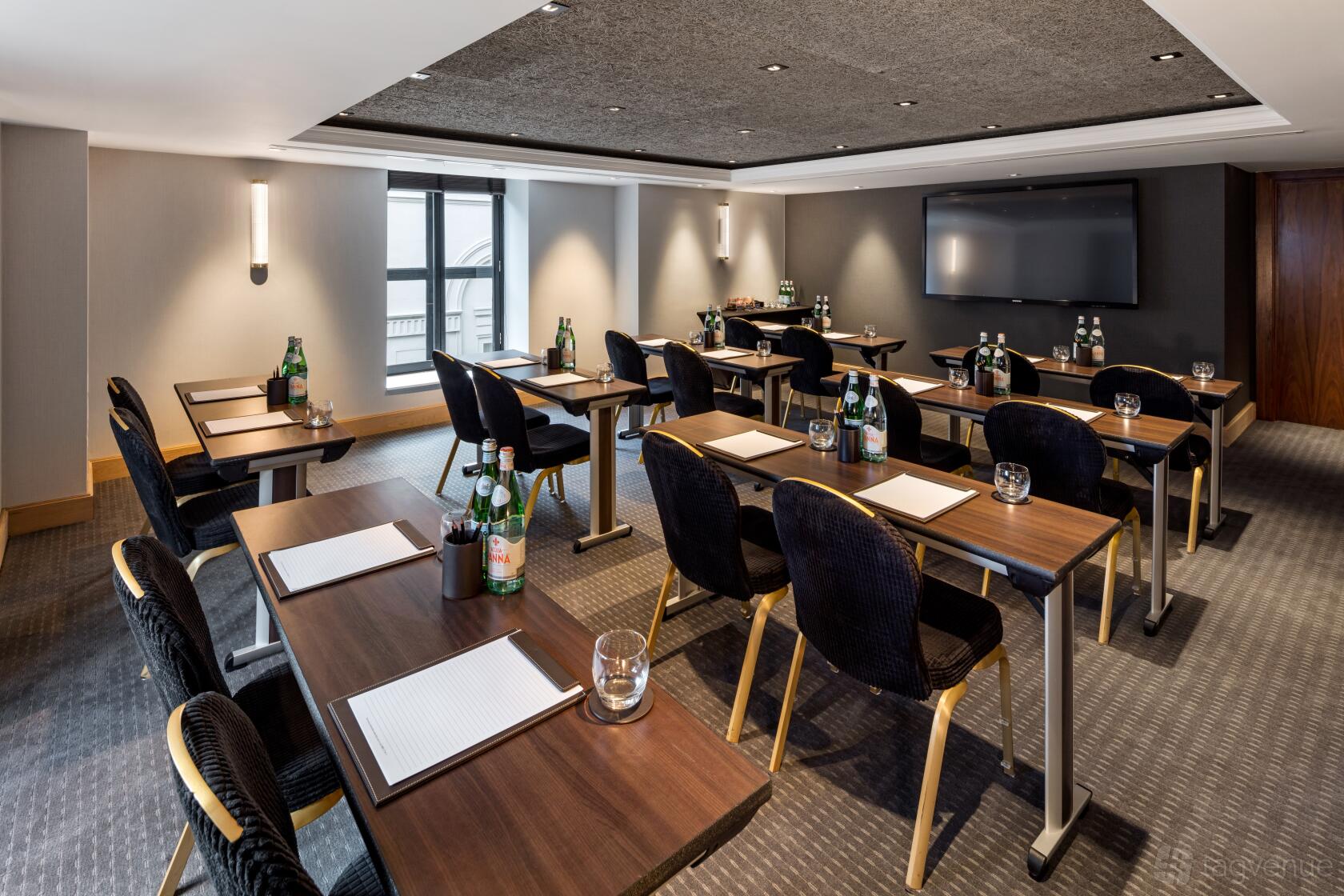 A hotel training room with rows of tables, black chairs, a wall-mounted screen, and bottled water at The Edwardian Manchester.