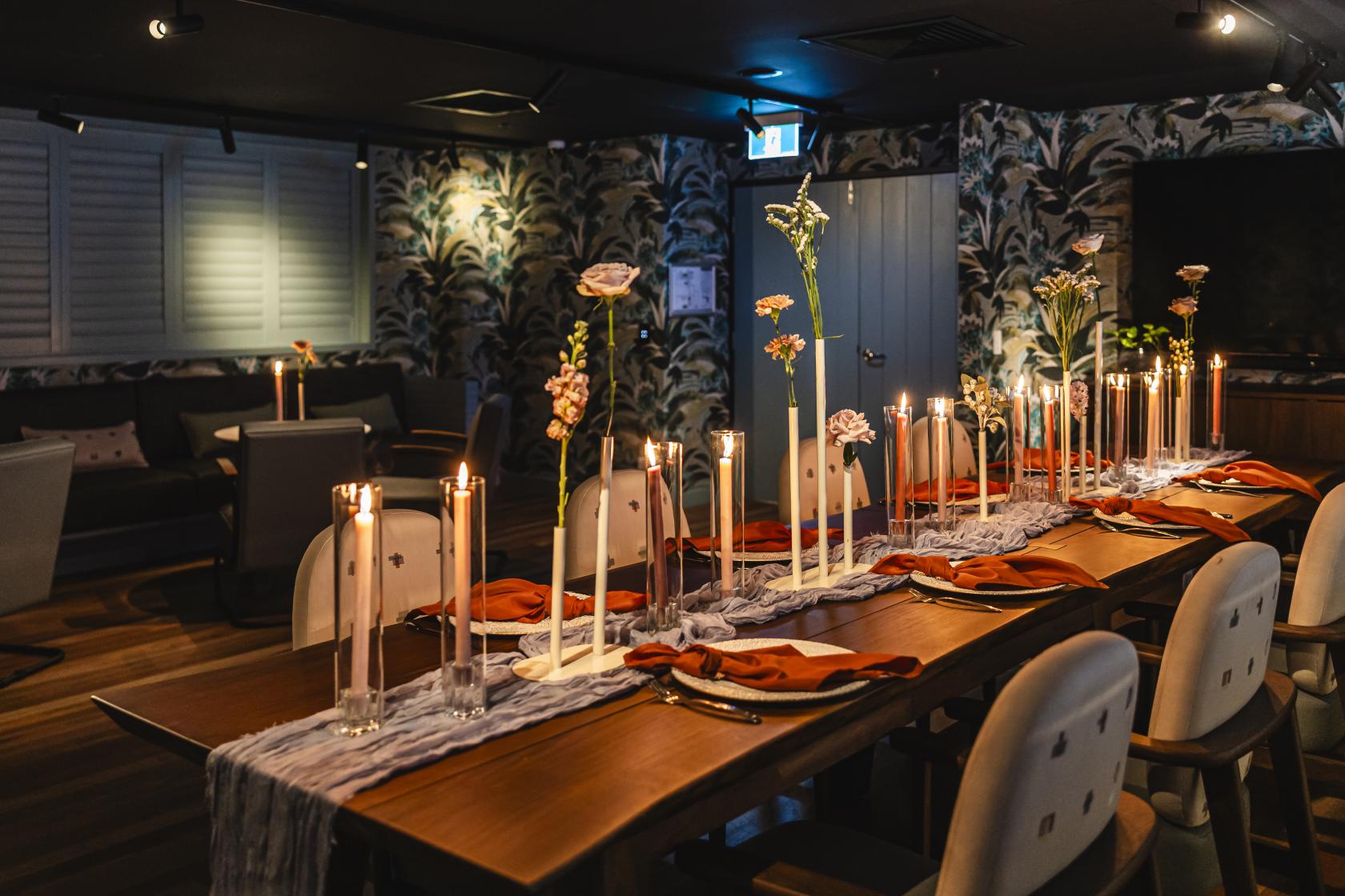A function room with a long wooden table, tall candles, single-stem flowers, and patterned wallpaper at Hotel Indigo Brisbane City Centre.
