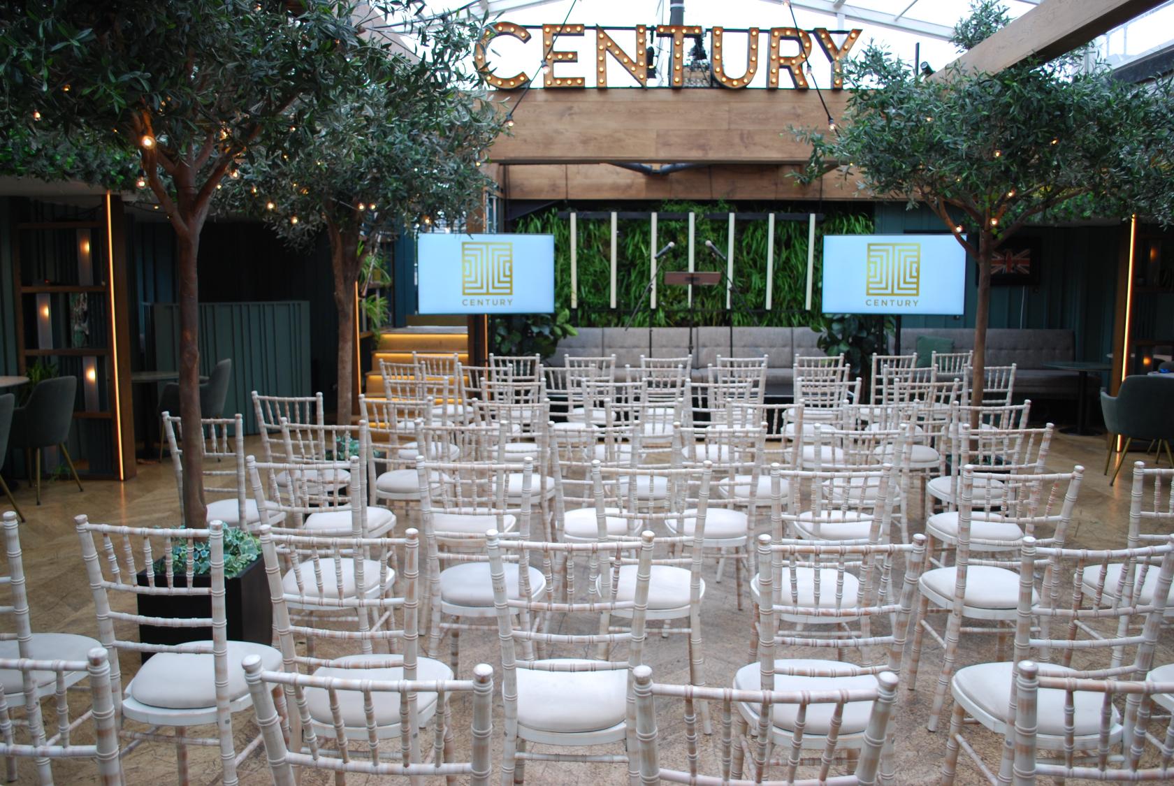 A rooftop terrace with rows of white chairs, potted trees, and large screens at Century Club.