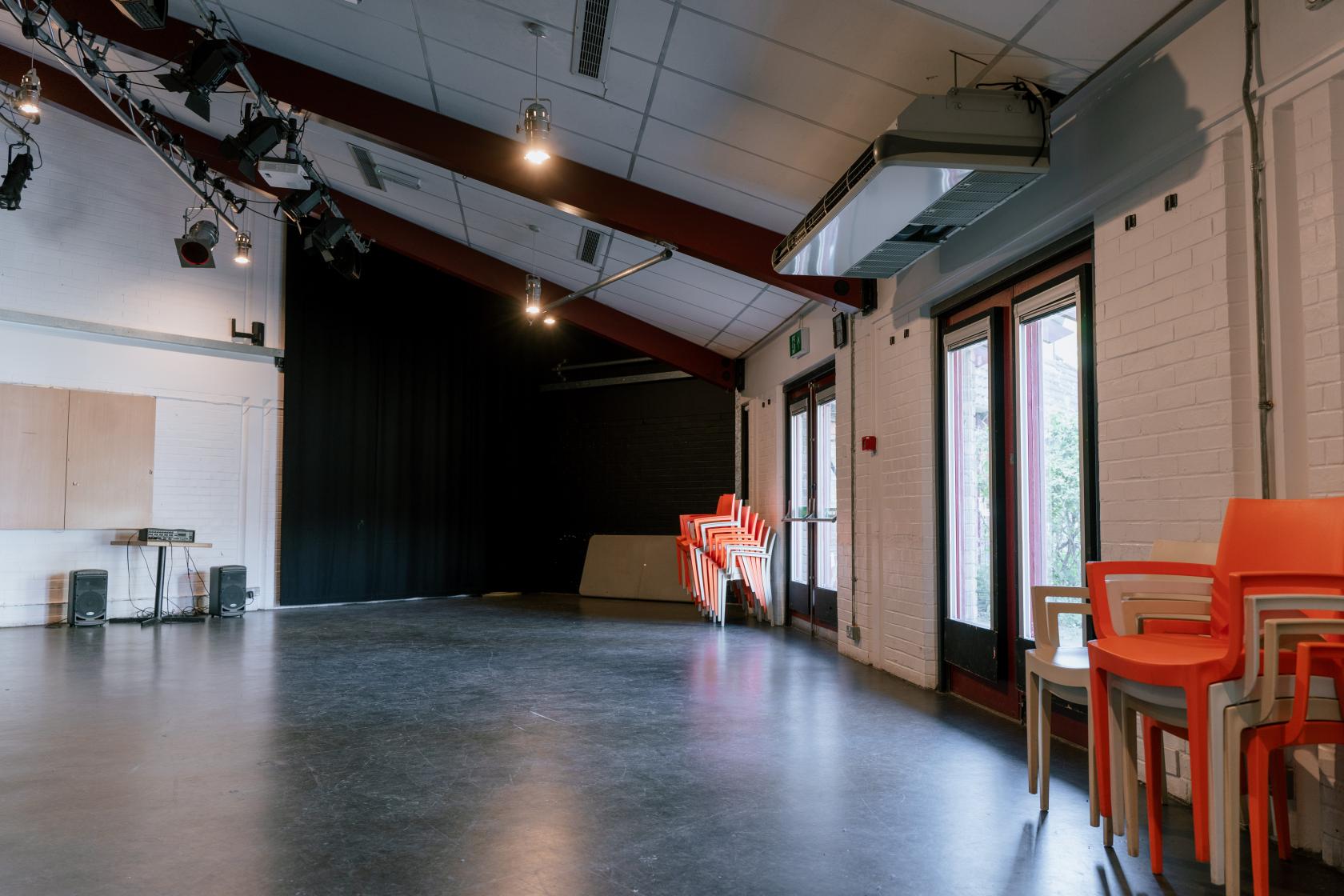 A community centre meeting room with a sloped ceiling, stage lighting, stacked orange chairs, and large windows at The Albany.
