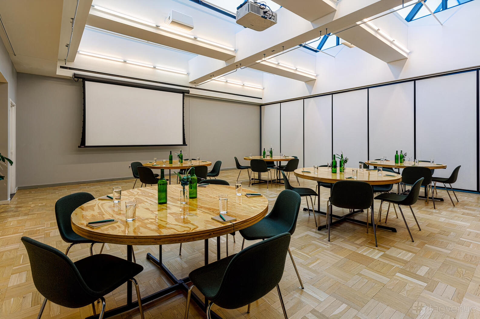 A meeting room with round wooden tables, black chairs, skylights, and a projector screen at The Emily Hotel.