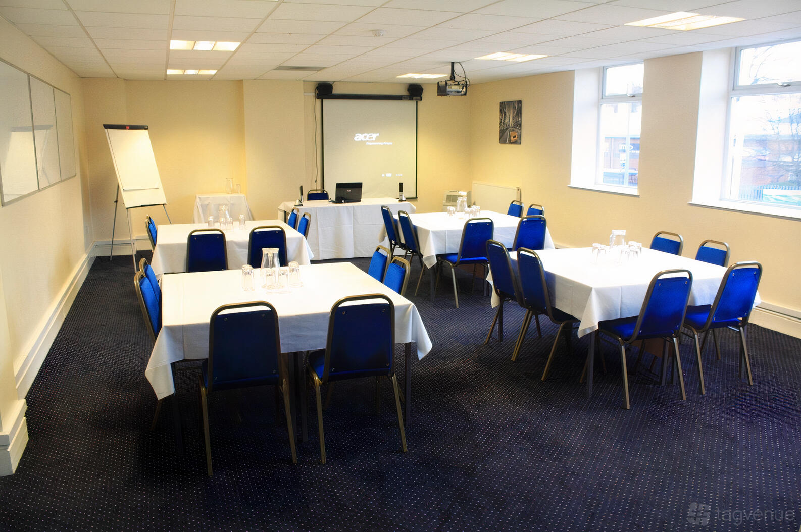 A meeting room with blue chairs, square tables covered with white cloths, a projector screen, and flipchart at The Saffron Centre.