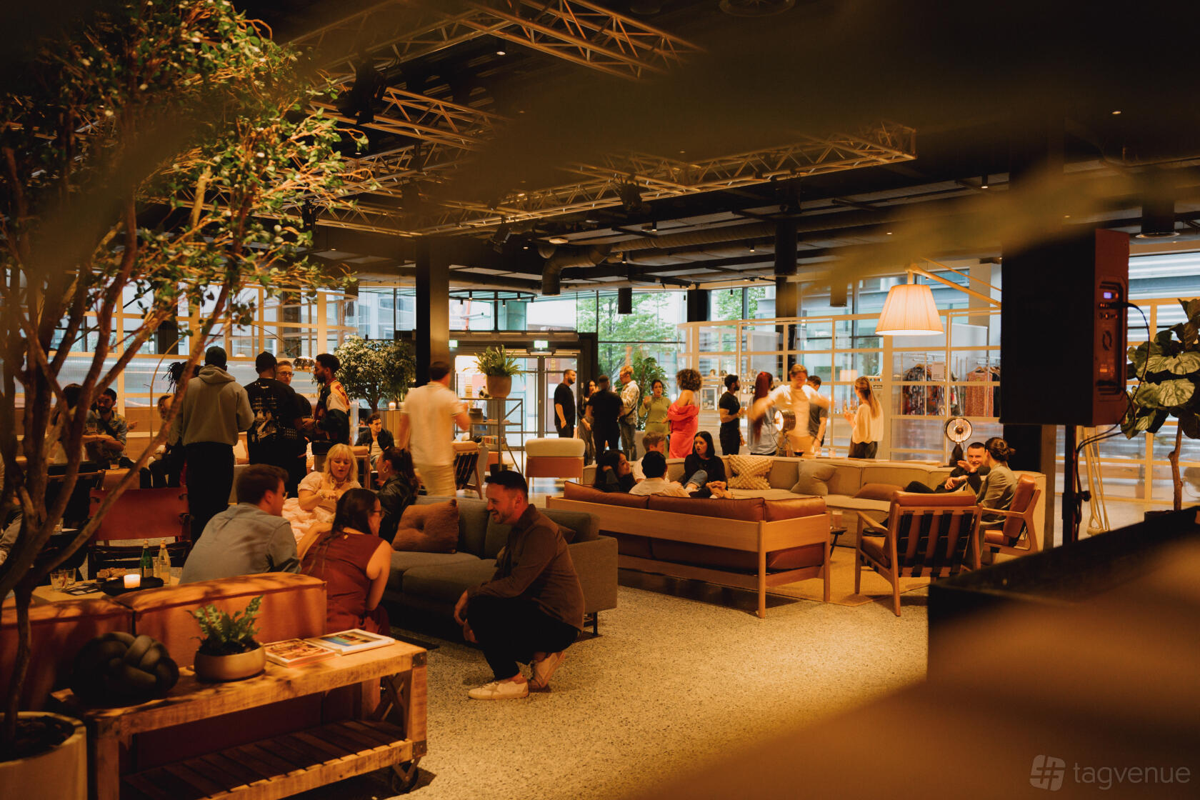 A lounge in a bar with floor-to-ceiling windows, indoor trees, and groups of people seated on sofas at Fearns Leeds Dock.
