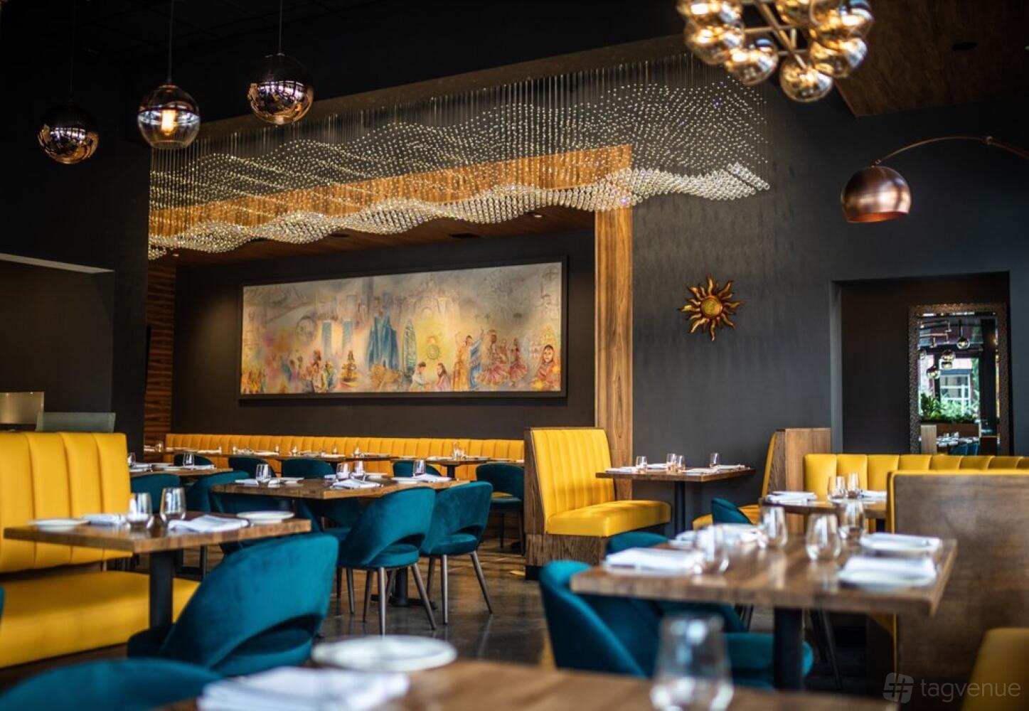 A restaurant with teal velvet chairs, yellow tufted booths, artistic wall decor, and modern pendant lighting at The Yellow Chilli Tustin.