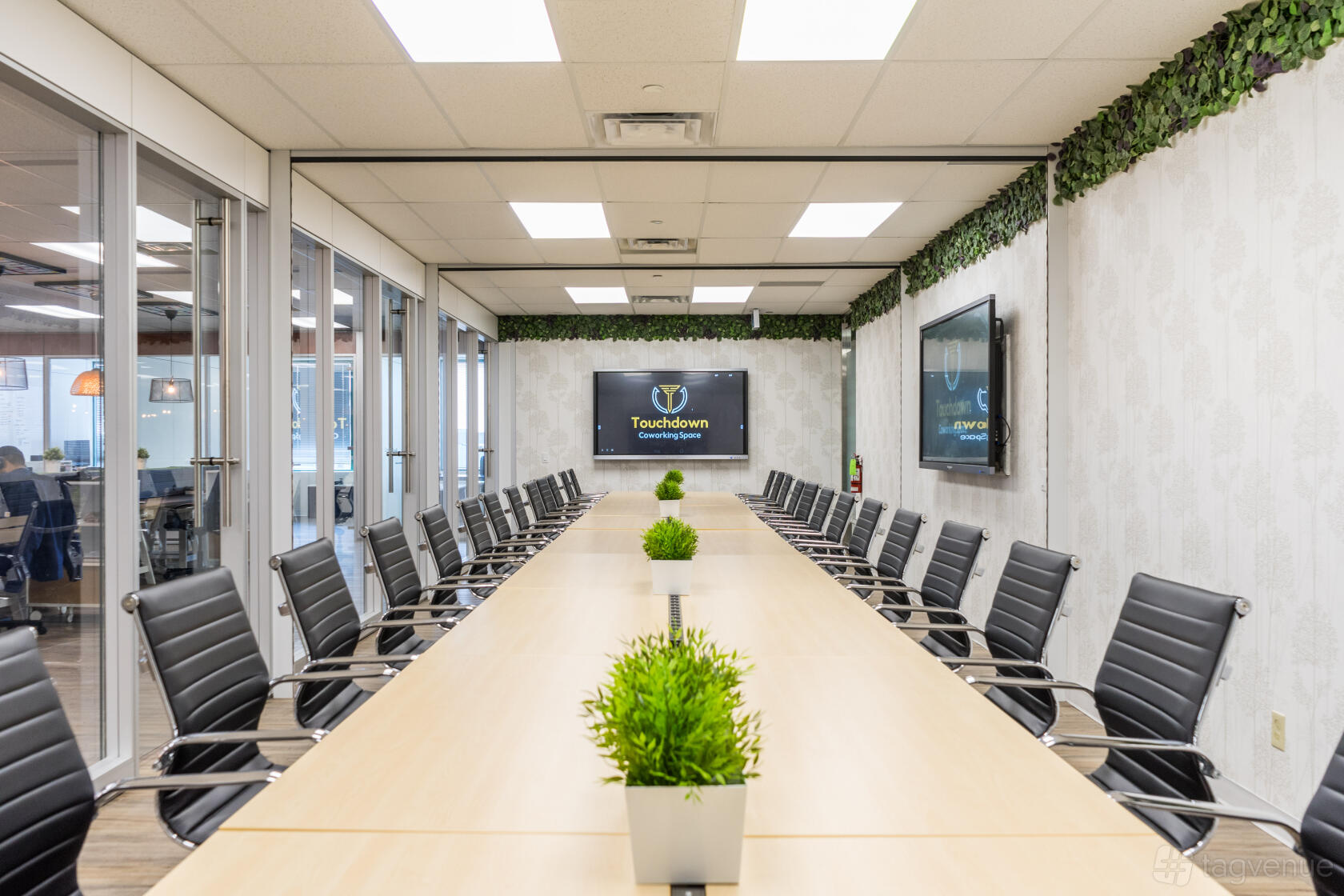 A coworking space with a long conference table, black chairs, potted plants, and wall-mounted screens at Touchdown Coworking.