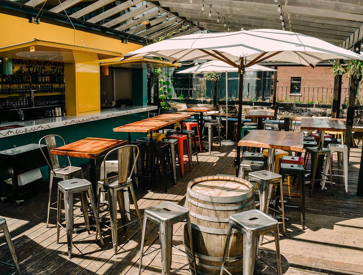 A pub terrace with wooden tables, metal stools, umbrellas, and an open bar counter at BAR HOP BREWCO.