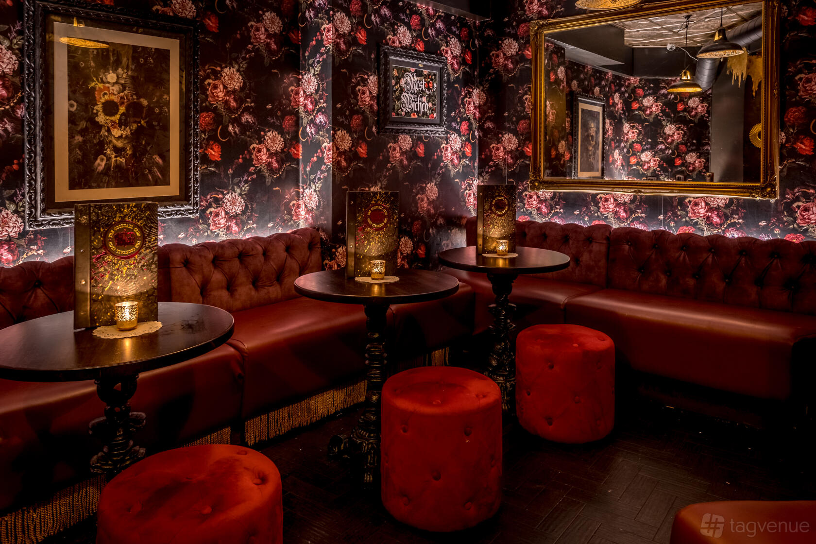 A cocktail bar with floral wallpaper, tufted red sofas, round ottomans, and candlelit tables at The Cocktail Club Shaftesbury Avenue.