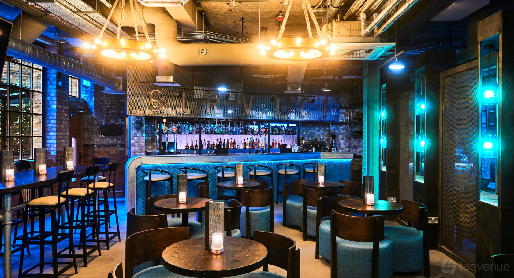 A hotel bar with exposed brick walls, blue accent lighting, and round tables with candles at New Road Hotel.
