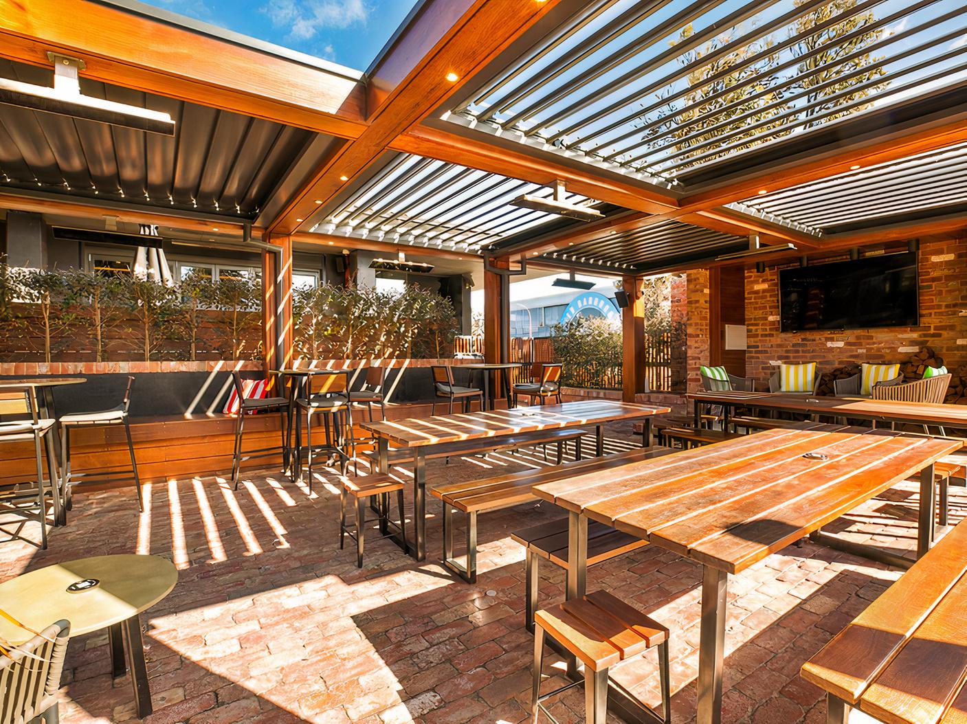 A terrace at a pub with wooden picnic tables, brick flooring, and a louvered pergola at The Cheeky Squire.