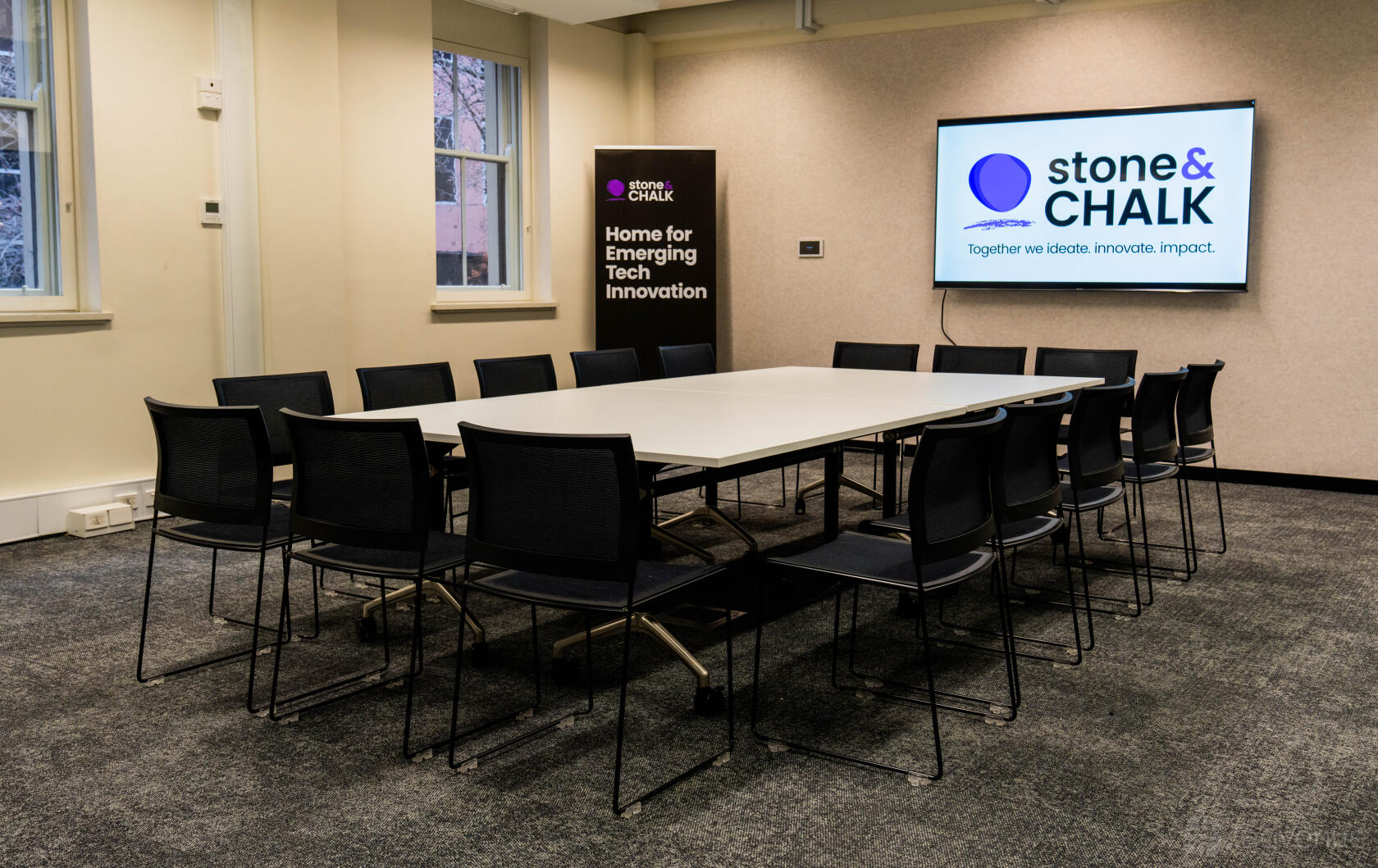 A meeting room with a long rectangular table, black chairs, wall-mounted screen, and high windows at Stone & Chalk Melbourne.