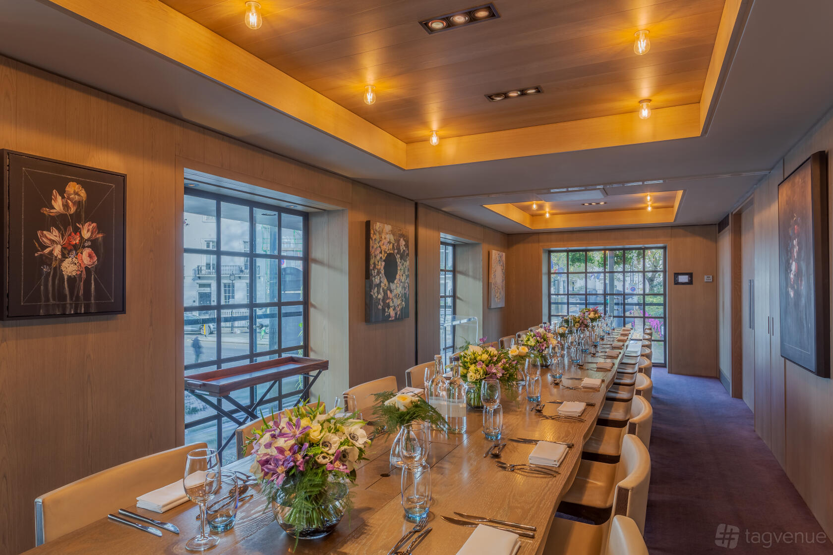An event space in a hotel with large windows, wood panel walls, floral artwork, and a long table with fresh flowers at The Hari