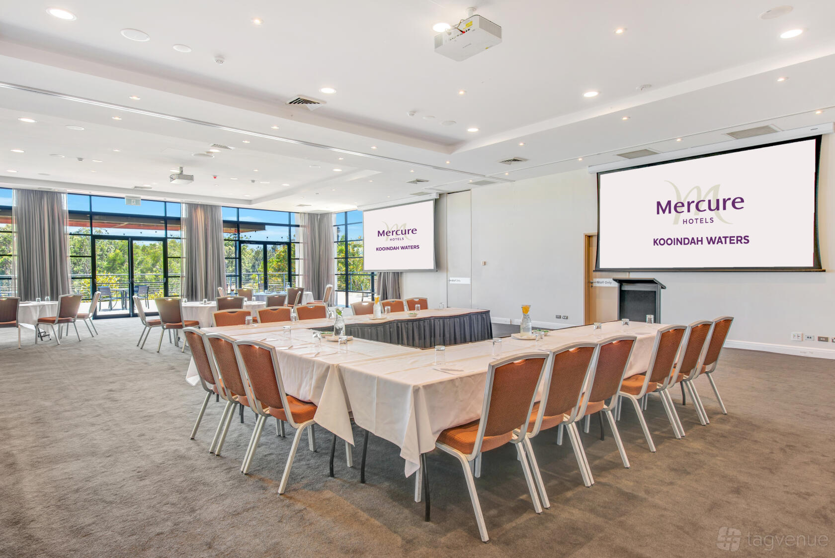A hotel meeting room with U-shaped tables, floor-to-ceiling windows, and two projector screens at Mercure Kooindah Waters.